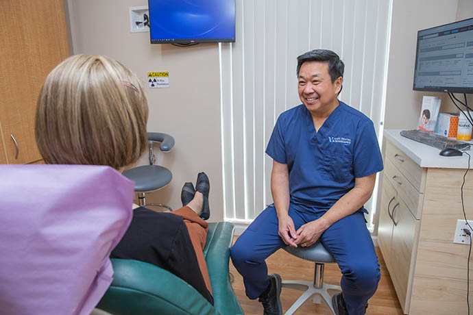 Dr. Doug Wong wearing a blue uniform seats in front of a lady patient inside his dental office
